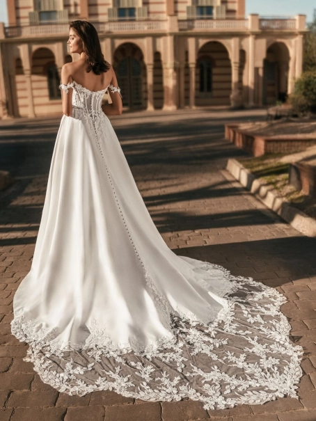 Blue by Enzoani Udell is a luxury satin ballgown wedding dress featuring a structured corset-inspired bodice, romantic off-the-shoulder straps, delicate floral appliqué detailing and a dramatic cathedral-length train. Designed for modern brides seeking a s