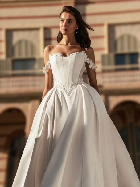 Blue by Enzoani Udell is a luxury satin ballgown wedding dress featuring a structured corset-inspired bodice, romantic off-the-shoulder straps, delicate floral appliqué detailing and a dramatic cathedral-length train. Designed for modern brides seeking a s