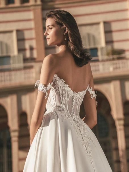 Blue by Enzoani Udell is a luxury satin ballgown wedding dress featuring a structured corset-inspired bodice, romantic off-the-shoulder straps, delicate floral appliqué detailing and a dramatic cathedral-length train. Designed for modern brides seeking a s