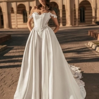 Blue by Enzoani Udell is a luxury satin ballgown wedding dress featuring a structured corset-inspired bodice, romantic off-the-shoulder straps, delicate floral appliqué detailing and a dramatic cathedral-length train. Designed for modern brides seeking a s