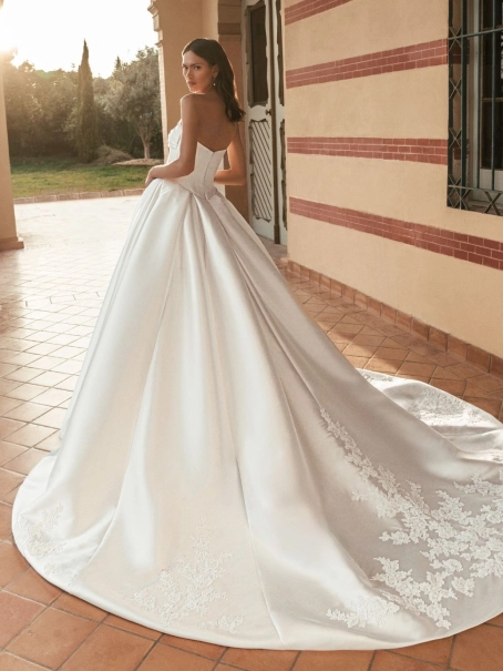 Blue by Enzoani Ulani is a luxury satin ballgown wedding dress featuring a structured corset-inspired bodice, elegant draped neckline, voluminous skirt with pockets and a dramatic cathedral-length train. Styled with a removable illusion lace high-neck topp