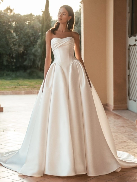 Blue by Enzoani Ulani is a luxury satin ballgown wedding dress featuring a structured corset-inspired bodice, elegant draped neckline, voluminous skirt with pockets and a dramatic cathedral-length train. Styled with a removable illusion lace high-neck topp
