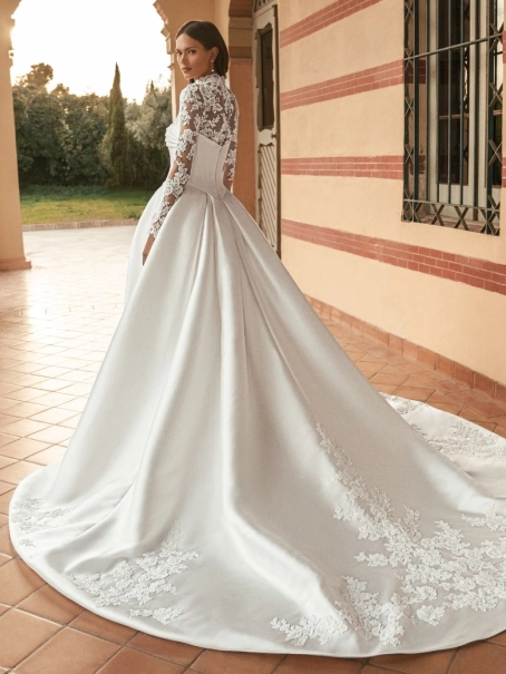Blue by Enzoani Ulani is a luxury satin ballgown wedding dress featuring a structured corset-inspired bodice, elegant draped neckline, voluminous skirt with pockets and a dramatic cathedral-length train. Styled with a removable illusion lace high-neck topp