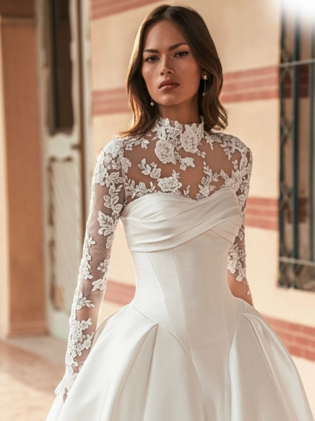 Blue by Enzoani Ulani is a luxury satin ballgown wedding dress featuring a structured corset-inspired bodice, elegant draped neckline, voluminous skirt with pockets and a dramatic cathedral-length train. Styled with a removable illusion lace high-neck topp