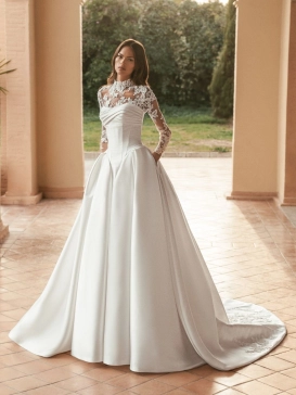 Blue by Enzoani Ulani is a luxury satin ballgown wedding dress featuring a structured corset-inspired bodice, elegant draped neckline, voluminous skirt with pockets and a dramatic cathedral-length train. Styled with a removable illusion lace high-neck topp