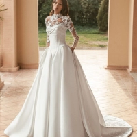 Blue by Enzoani Ulani is a luxury satin ballgown wedding dress featuring a structured corset-inspired bodice, elegant draped neckline, voluminous skirt with pockets and a dramatic cathedral-length train. Styled with a removable illusion lace high-neck topp