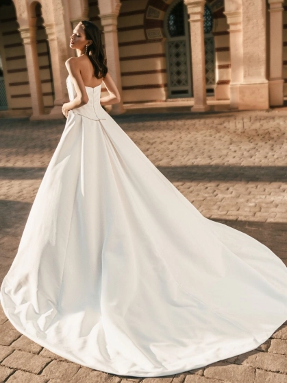 Blue by Enzoani Uleana is a luxury wedding dress featuring a structured satin corset bodice with detachable crystal bead draping, a shimmering fitted column silhouette and a dramatic detachable overskirt with sweeping train. Designed for brides seeking a v