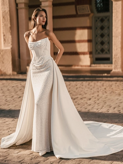 Blue by Enzoani Uleana is a luxury wedding dress featuring a structured satin corset bodice with detachable crystal bead draping, a shimmering fitted column silhouette and a dramatic detachable overskirt with sweeping train. Designed for brides seeking a v
