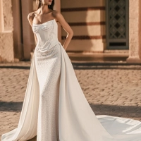 Blue by Enzoani Uleana is a luxury wedding dress featuring a structured satin corset bodice with detachable crystal bead draping, a shimmering fitted column silhouette and a dramatic detachable overskirt with sweeping train. Designed for brides seeking a v