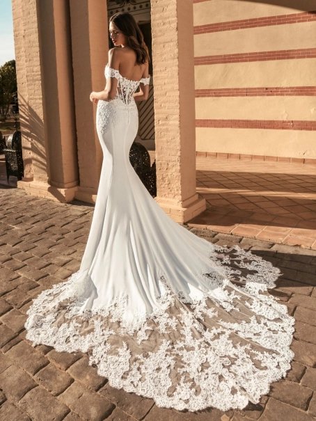 Blue by Enzoani Umani is a luxury fitted mermaid wedding dress featuring a romantic off-the-shoulder neckline, sculpted corset-inspired bodice, illusion side detailing and delicate floral lace appliqués. Designed for brides seeking a sophisticated designer