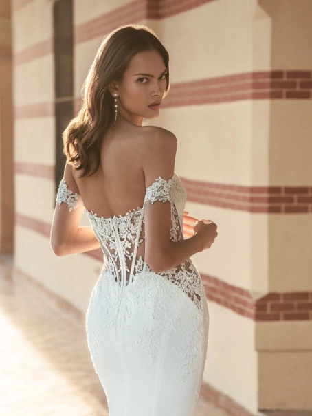 Blue by Enzoani Umani is a luxury fitted mermaid wedding dress featuring a romantic off-the-shoulder neckline, sculpted corset-inspired bodice, illusion side detailing and delicate floral lace appliqués. Designed for brides seeking a sophisticated designer