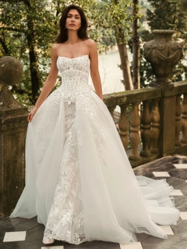 Bride wearing the Enzoani WALINA wedding dress featuring a strapless lace corset bodice, fitted floral lace silhouette and detachable tulle overskirt on an elegant lakeside terrace.