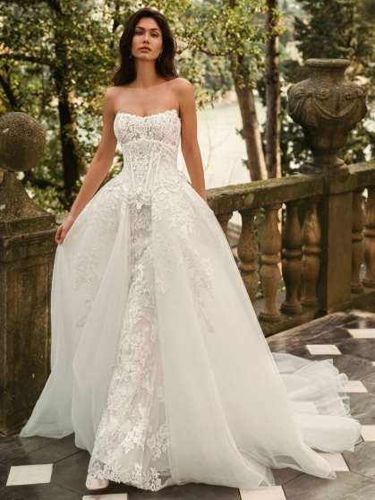 Bride wearing the Enzoani WALINA wedding dress featuring a strapless lace corset bodice, fitted floral lace silhouette and detachable tulle overskirt on an elegant lakeside terrace.