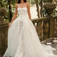 Bride wearing the Enzoani WALINA wedding dress featuring a strapless lace corset bodice, fitted floral lace silhouette and detachable tulle overskirt on an elegant lakeside terrace.
