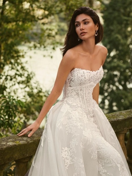 Bride wearing the Enzoani WALINA wedding dress featuring a strapless lace corset bodice, fitted floral lace silhouette and detachable tulle overskirt on an elegant lakeside terrace.