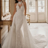 Enzoani Wednesday luxury wedding dress with a fitted mermaid silhouette, illusion halter neckline, floral lace embroidery, sculpted corset bodice, detachable overskirt, and dramatic cathedral train. Couture bridal gown by Enzoani combining modern glamour,