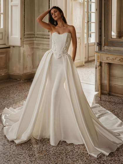 Enzoani Wilomina is a luxury designer wedding dress featuring a structured corset-inspired bodice, elegant satin construction, floral appliqué detailing and a dramatic detachable overskirt with cathedral-length train. Combining modern bridal fashion with t