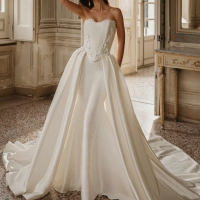 Enzoani Wilomina is a luxury designer wedding dress featuring a structured corset-inspired bodice, elegant satin construction, floral appliqué detailing and a dramatic detachable overskirt with cathedral-length train. Combining modern bridal fashion with t