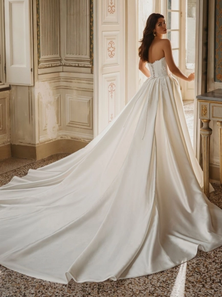 Enzoani Wilomina is a luxury designer wedding dress featuring a structured corset-inspired bodice, elegant satin construction, floral appliqué detailing and a dramatic detachable overskirt with cathedral-length train. Combining modern bridal fashion with t