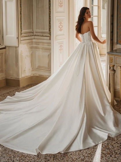 Enzoani Wilomina is a luxury designer wedding dress featuring a structured corset-inspired bodice, elegant satin construction, floral appliqué detailing and a dramatic detachable overskirt with cathedral-length train. Combining modern bridal fashion with t