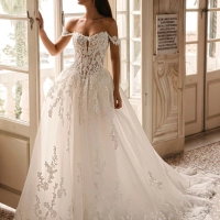 Enzoani Winona is a luxury ballgown wedding dress featuring an illusion corset bodice, romantic off-the-shoulder straps, intricate floral lace appliqué and a dramatic cathedral-length train. Designed for brides seeking a princess wedding dress with couture