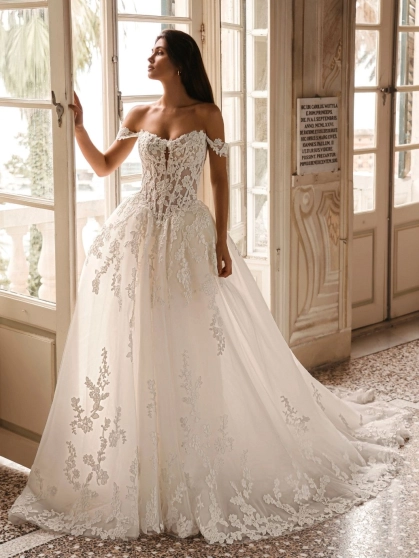 Enzoani Winona is a luxury ballgown wedding dress featuring an illusion corset bodice, romantic off-the-shoulder straps, intricate floral lace appliqué and a dramatic cathedral-length train. Designed for brides seeking a princess wedding dress with couture