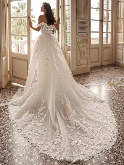 Enzoani Winona is a luxury ballgown wedding dress featuring an illusion corset bodice, romantic off-the-shoulder straps, intricate floral lace appliqué and a dramatic cathedral-length train. Designed for brides seeking a princess wedding dress with couture