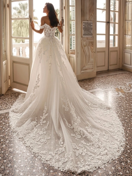 Enzoani Winona is a luxury ballgown wedding dress featuring an illusion corset bodice, romantic off-the-shoulder straps, intricate floral lace appliqué and a dramatic cathedral-length train. Designed for brides seeking a princess wedding dress with couture