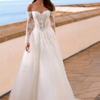 Love Faiza is a romantic A-line wedding dress from the 2027 collection featuring an illusion lace corset bodice, detachable off-the-shoulder long lace sleeves and a flowing soft tulle skirt with floral lace appliqués. Designed by Enzoani for brides seeking