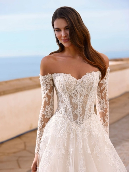 Love Faiza is a romantic A-line wedding dress from the 2027 collection featuring an illusion lace corset bodice, detachable off-the-shoulder long lace sleeves and a flowing soft tulle skirt with floral lace appliqués. Designed by Enzoani for brides seeking