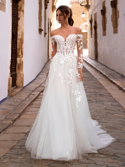 Love by Enzoani FISELLE is a romantic A-line wedding dress featuring a sheer illusion corset bodice adorned with dimensional floral lace appliqués that cascade into a soft layered tulle skirt. The gown is completed with detachable off-the-shoulder illusion