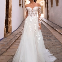 Love by Enzoani FISELLE is a romantic A-line wedding dress featuring a sheer illusion corset bodice adorned with dimensional floral lace appliqués that cascade into a soft layered tulle skirt. The gown is completed with detachable off-the-shoulder illusion