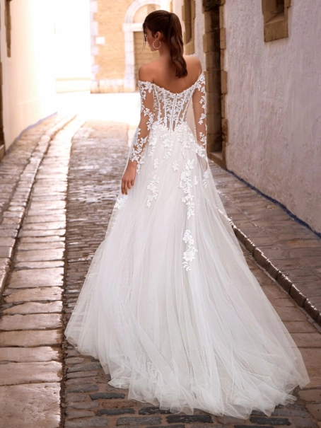 Love by Enzoani FISELLE is a romantic A-line wedding dress featuring a sheer illusion corset bodice adorned with dimensional floral lace appliqués that cascade into a soft layered tulle skirt. The gown is completed with detachable off-the-shoulder illusion