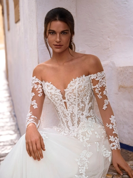 Love by Enzoani FISELLE is a romantic A-line wedding dress featuring a sheer illusion corset bodice adorned with dimensional floral lace appliqués that cascade into a soft layered tulle skirt. The gown is completed with detachable off-the-shoulder illusion