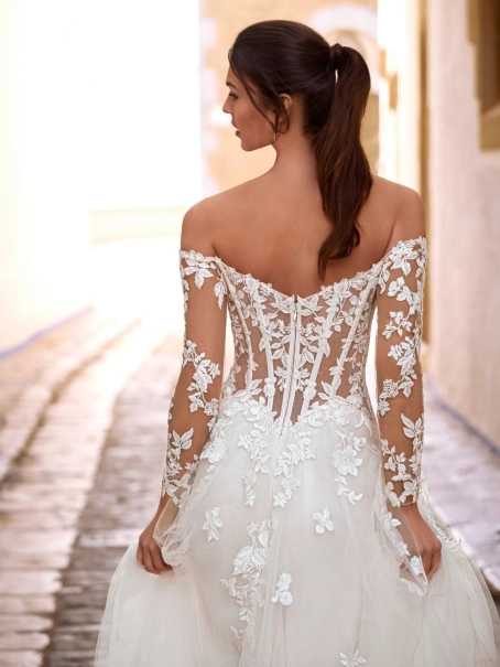 Love by Enzoani FISELLE is a romantic A-line wedding dress featuring a sheer illusion corset bodice adorned with dimensional floral lace appliqués that cascade into a soft layered tulle skirt. The gown is completed with detachable off-the-shoulder illusion