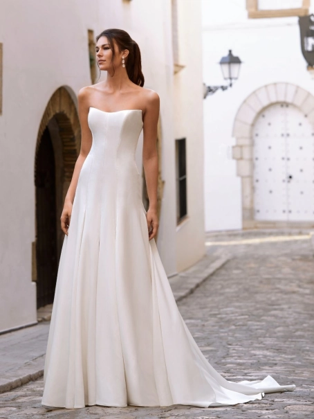 Love by Enzoani FLORENZA is a sophisticated fit and flare wedding dress crafted from luxurious satin with clean, sculptural lines. The gown features an elegant attached draped off-the-shoulder shawl, complemented by sheer illusion floral lace long sleeves