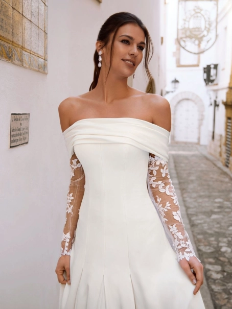 Love by Enzoani FLORENZA is a sophisticated fit and flare wedding dress crafted from luxurious satin with clean, sculptural lines. The gown features an elegant attached draped off-the-shoulder shawl, complemented by sheer illusion floral lace long sleeves
