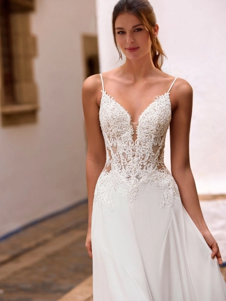 Love by Enzoani FLORETTA is an effortlessly romantic fit and flare wedding dress featuring a sheer illusion bodice adorned with intricate floral lace appliqués and delicate spaghetti straps. Soft illusion side panels create a flattering, sculpted silhouett