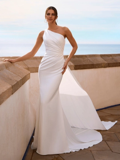 Love by Enzoani FLYNNE is a contemporary minimalist wedding dress crafted in luxurious crepe. This elegant fit and flare silhouette features a softly draped one-shoulder neckline, figure-enhancing ruching across the bodice, and a striking open back framed