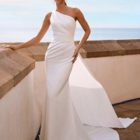 Love by Enzoani FLYNNE is a contemporary minimalist wedding dress crafted in luxurious crepe. This elegant fit and flare silhouette features a softly draped one-shoulder neckline, figure-enhancing ruching across the bodice, and a striking open back framed