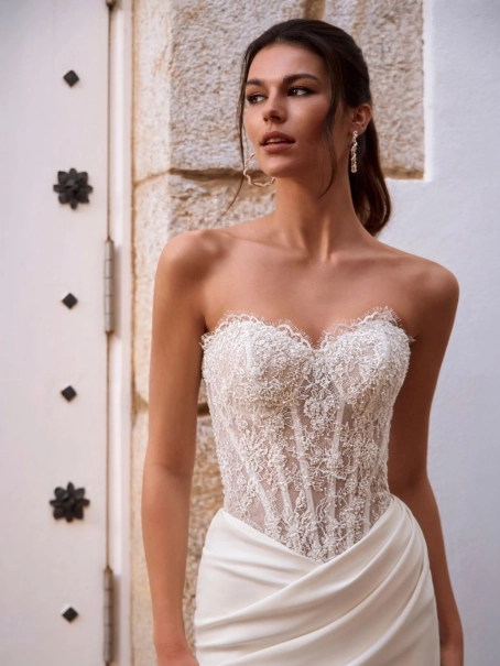 Love by Enzoani FOSIRA is a glamorous fit and flare wedding dress that combines a sparkling beaded illusion corset bodice with luxurious crepe. Featuring a romantic sweetheart neckline, exposed corset boning, intricate beadwork and soft draping across the