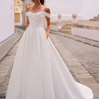 Love by Enzoani FRANCINA is a timeless satin ball gown wedding dress featuring a softly draped off-the-shoulder neckline, flattering ruched bodice and a full pleated skirt with hidden pockets. Combining classic bridal elegance with modern functionality, th