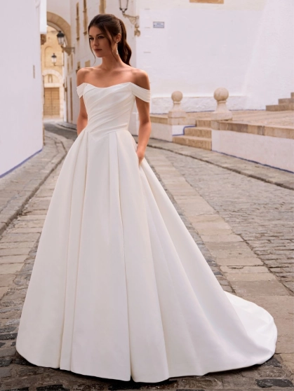 Love by Enzoani FRANCINA is a timeless satin ball gown wedding dress featuring a softly draped off-the-shoulder neckline, flattering ruched bodice and a full pleated skirt with hidden pockets. Combining classic bridal elegance with modern functionality, th