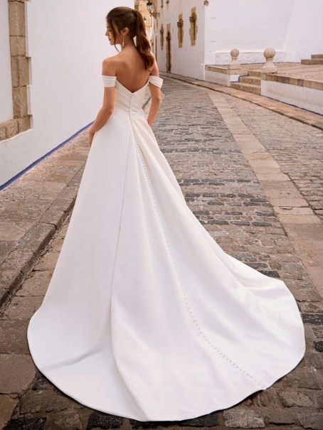 Love by Enzoani FRANCINA is a timeless satin ball gown wedding dress featuring a softly draped off-the-shoulder neckline, flattering ruched bodice and a full pleated skirt with hidden pockets. Combining classic bridal elegance with modern functionality, th