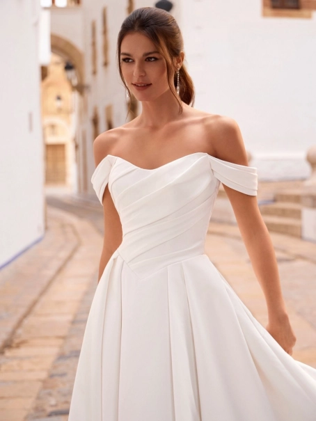 Love by Enzoani FRANCINA is a timeless satin ball gown wedding dress featuring a softly draped off-the-shoulder neckline, flattering ruched bodice and a full pleated skirt with hidden pockets. Combining classic bridal elegance with modern functionality, th