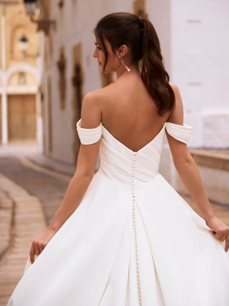 Love by Enzoani FRANCINA is a timeless satin ball gown wedding dress featuring a softly draped off-the-shoulder neckline, flattering ruched bodice and a full pleated skirt with hidden pockets. Combining classic bridal elegance with modern functionality, th